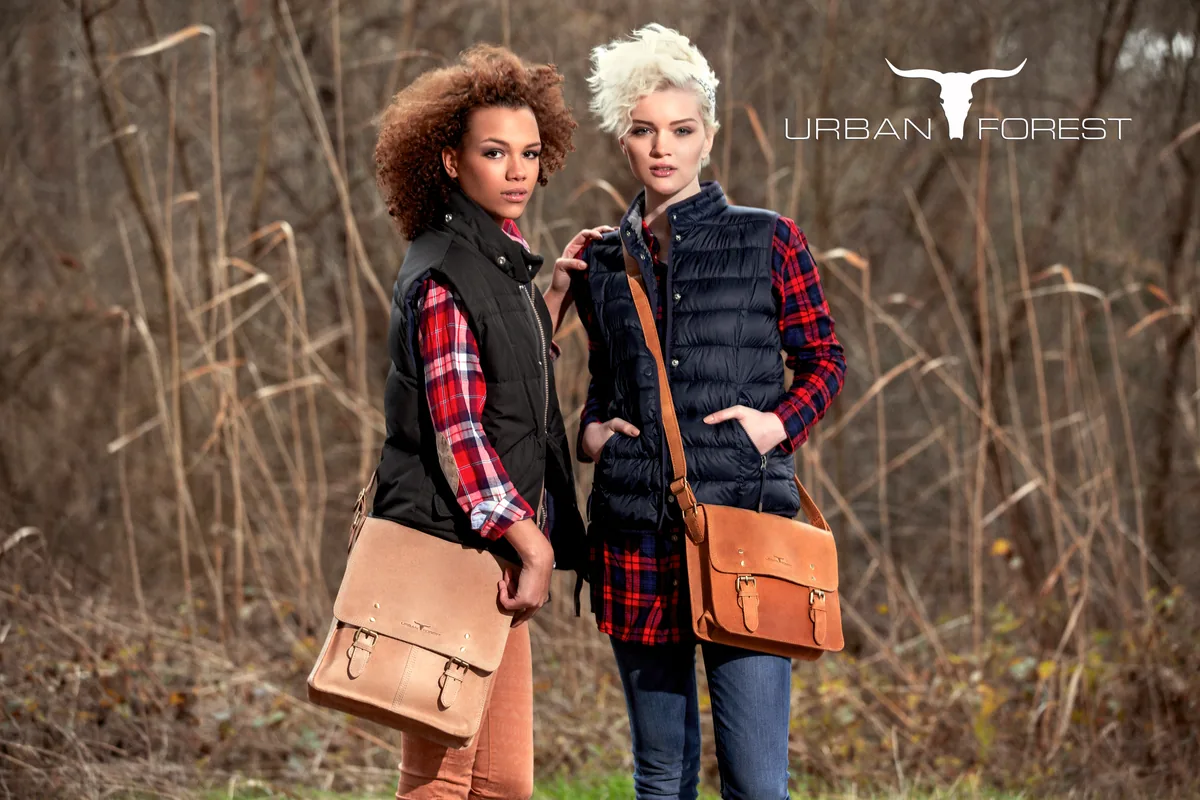 Zwei Frauen in Westen und karierten Hemden stehen vor trockenem Schilf, jede mit einer braunen Umhängetasche. Oben rechts im Bild steht das URBAN-FOREST-Logo mit Stierkopf.