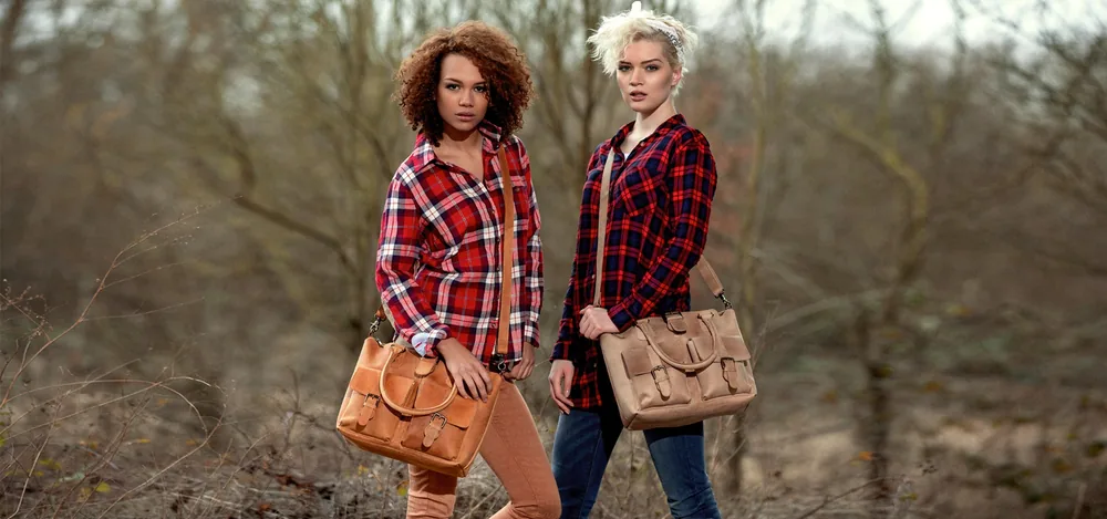 Zwei Frauen in karierten Hemden stehen in einem kahlen Wald, jede mit einer braunen Tasche.