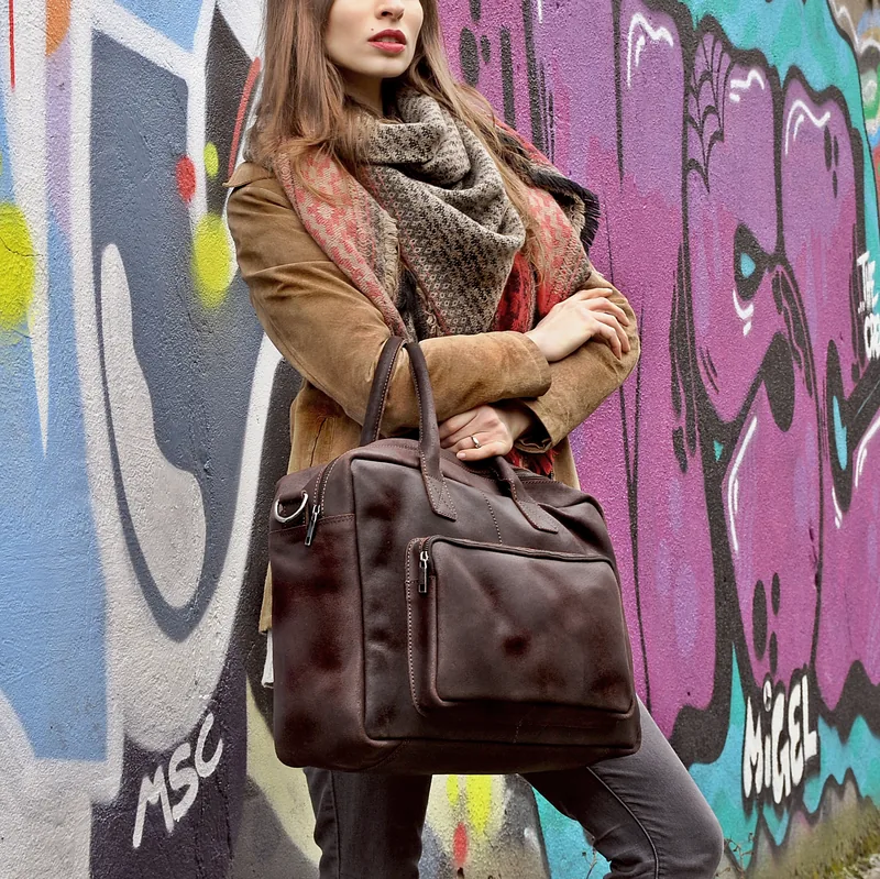 Frau in beiger Jacke und gemustertem Schal lehnt an einer bunt bemalten Graffiti-Wand und hält eine dunkelbraune Tasche.