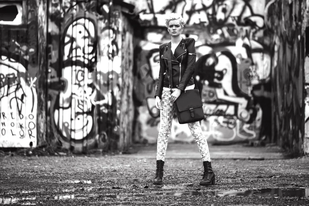 Schwarzweißfoto: Frau in dunkler Jacke und gemusterter Hose steht breitbeinig auf einem Platz vor Graffiti-Wänden, eine dunkle Tasche in der Hand.