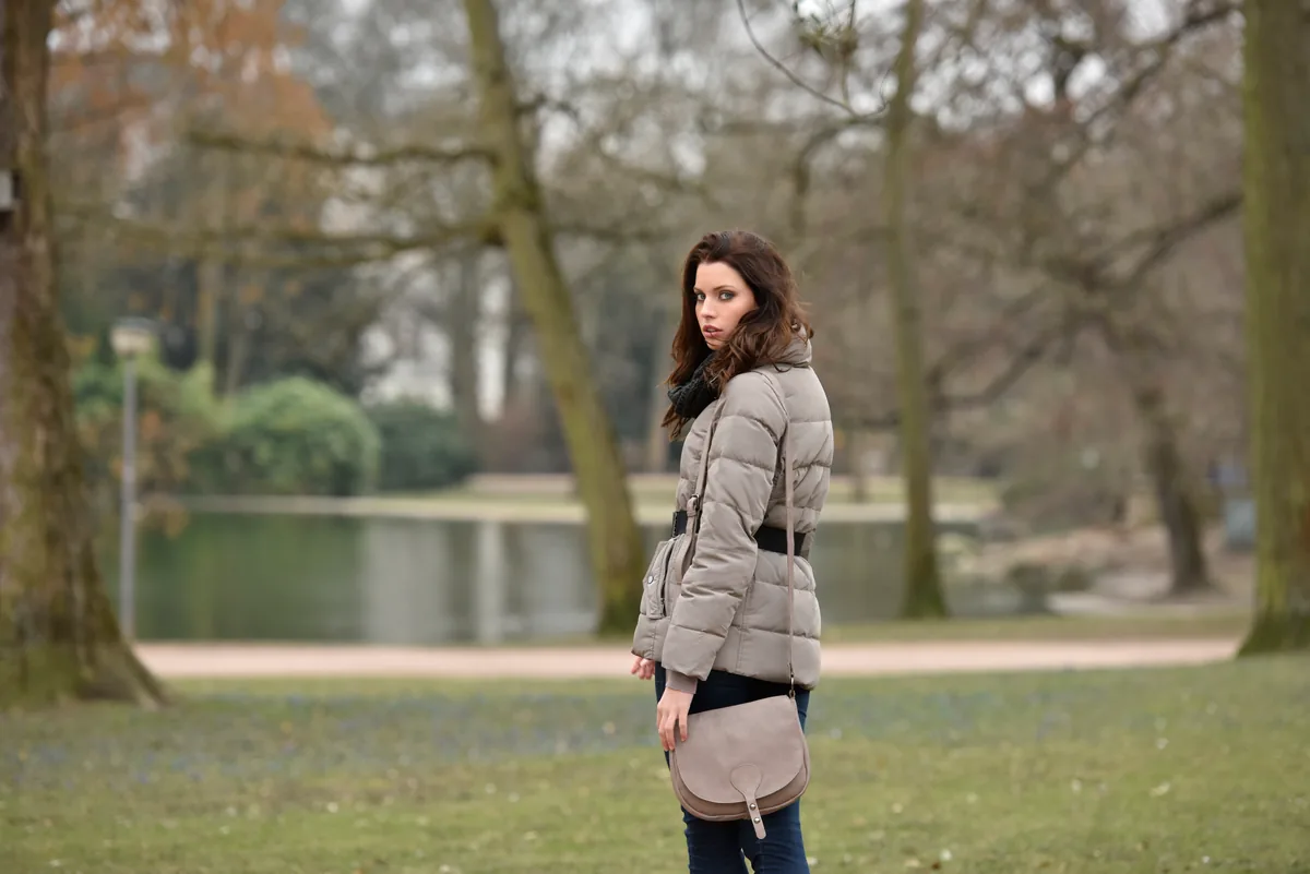 Frau in grauer Daunenjacke blickt über die Schulter, hinter ihr ein Parkteich und kahle Bäume, an der Hüfte eine graue Umhängetasche.