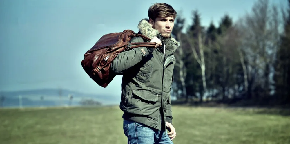 Mann in grüner Jacke trägt eine braune Reisetasche über der Schulter, im Hintergrund Wiese und Bäume.