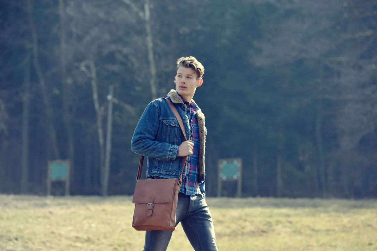 Mann in Jeansjacke und kariertem Hemd geht über eine Wiese, hinter ihm ein dunkler Wald. Er trägt eine braune Umhängetasche.