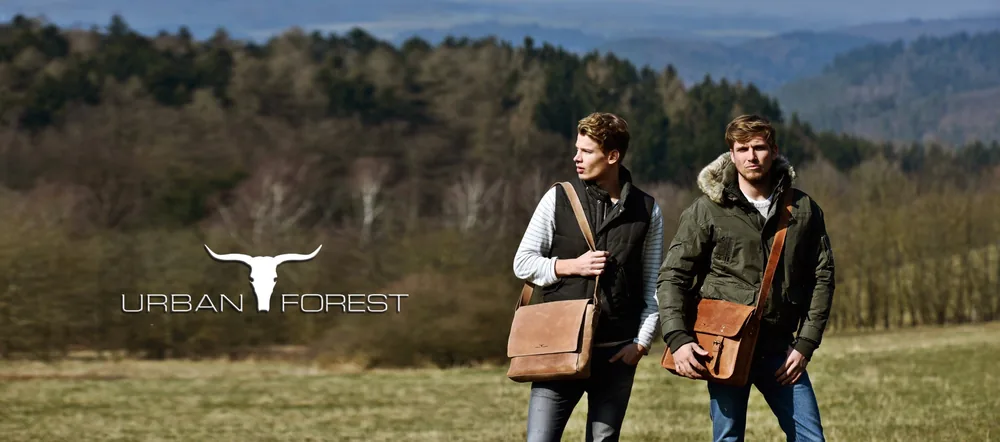 Zwei Männer gehen über eine Wiese, hinter ihnen Wald und Hügel, beide tragen eine braune Umhängetasche. Links im Bild steht das URBAN-FOREST-Logo mit Stierkopf.